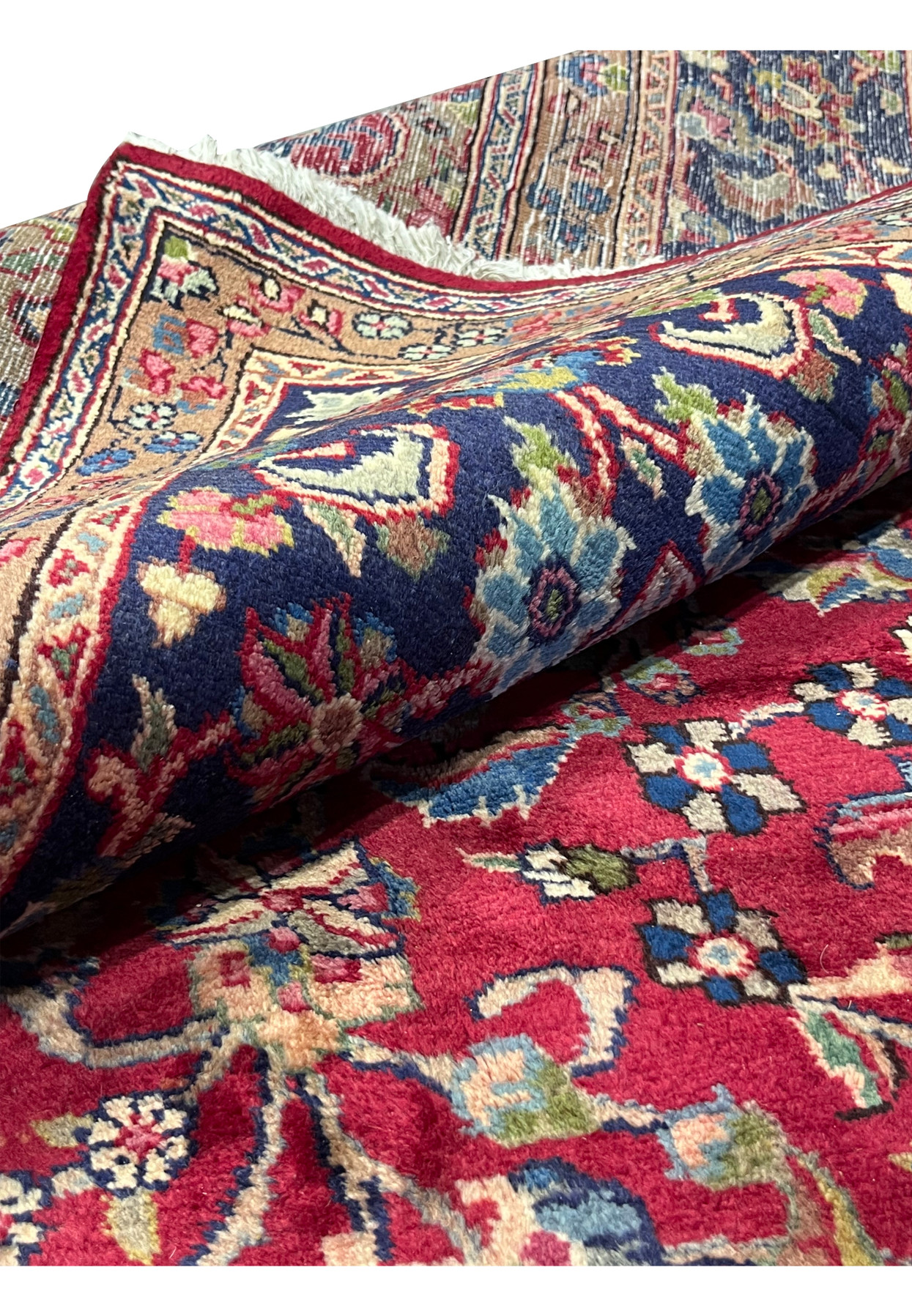 Rolled corner of 10x13 Persian Mashad rug, navy floral border framing cherry red field, full thick wool pile