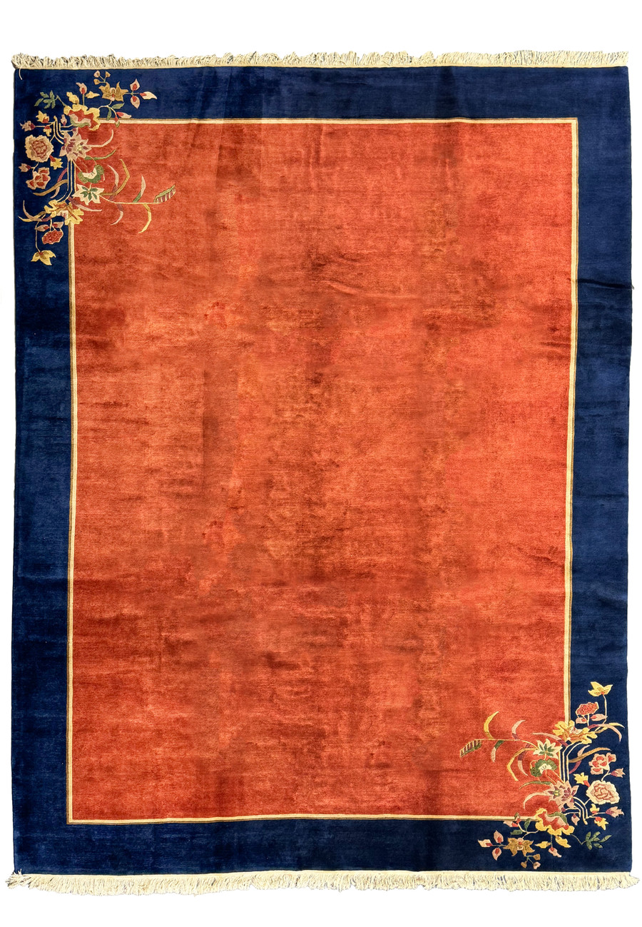 A top-down view of a large, 10x13 modern Oriental Art Deco rug with a central burnt orange field, bordered by deep navy blue with a golden edge, and a corner floral design in gold, blue, and pink tones.