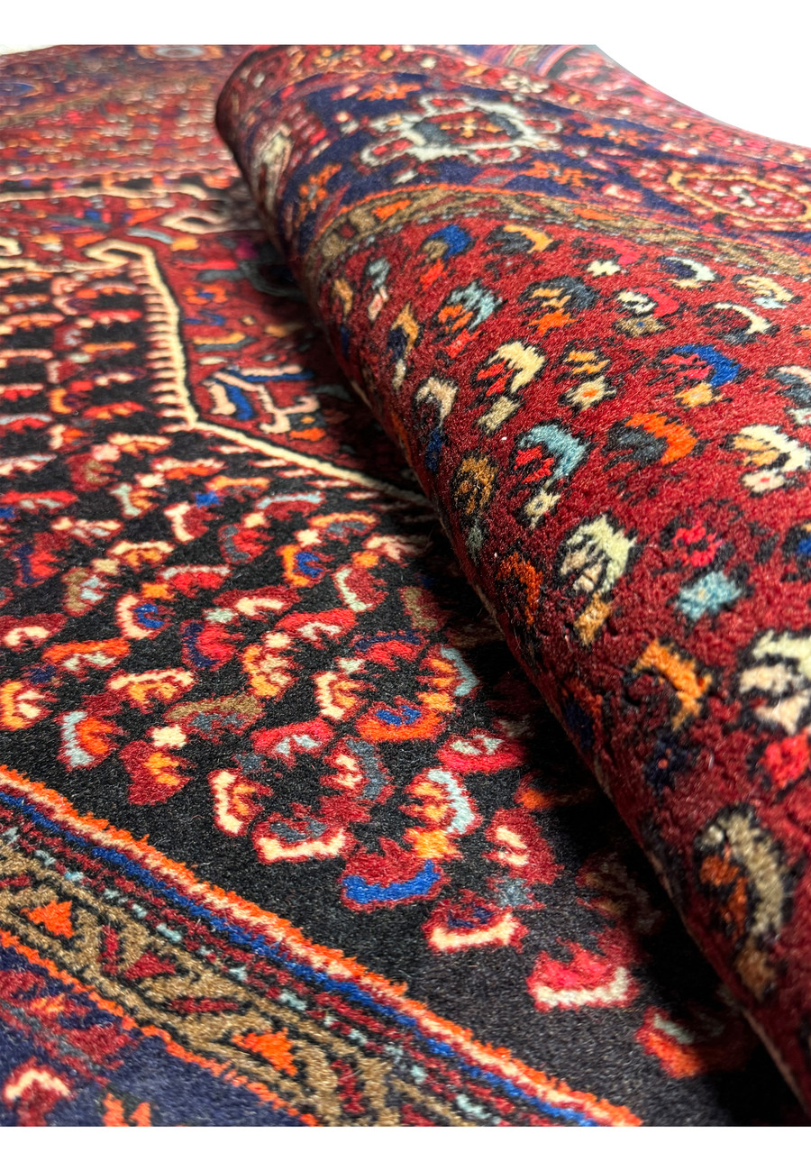 Perspective view of a Persian Gholtogh rug rolled slightly to showcase the thickness and texture of the weave, with vivid red and blue patterns visible
