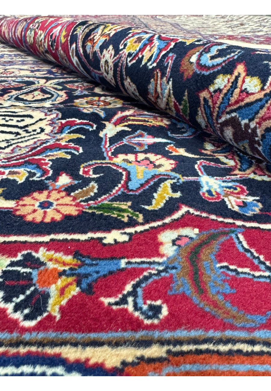 Close-up view of a Persian Mashad rug partially rolled, highlighting vibrant floral patterns and intricate motifs in deep red, blue, and ivory, showcasing the rich colors and detailed craftsmanship typical of traditional Persian rugs