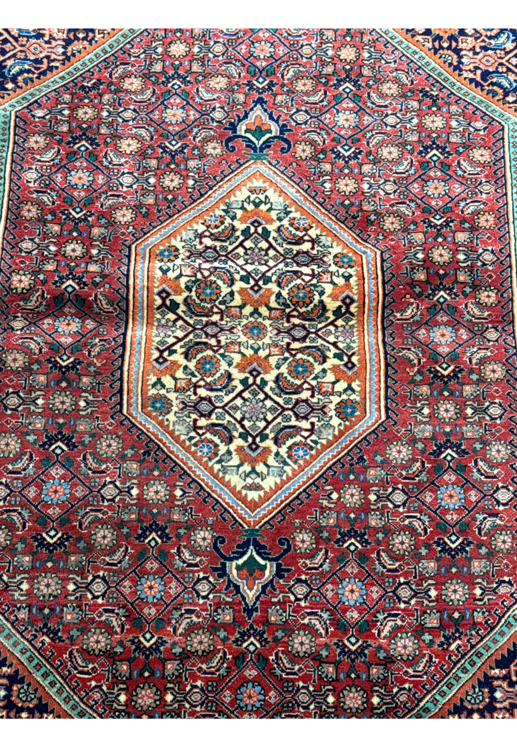 A detailed image of a Persian Bijar rug laid flat, showcasing the central medallion design with a red background and intricate floral patterns in indigo, ivory, and orange hues