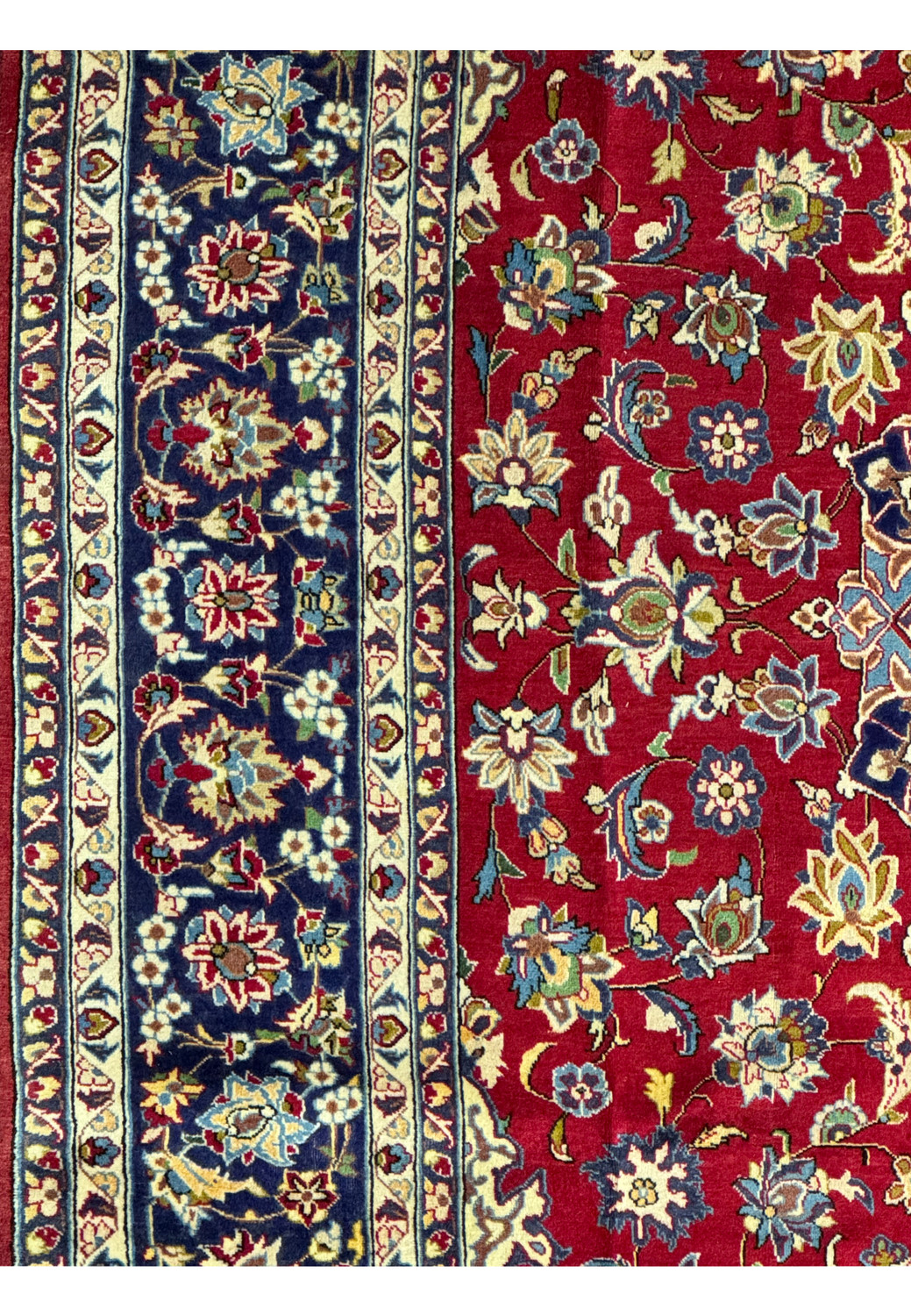 A zoomed-in view of the Persian Isfahan rug's edge, displaying the intricate border patterns with reciprocal trefoil designs in white, blue, and gold, against a dark navy background