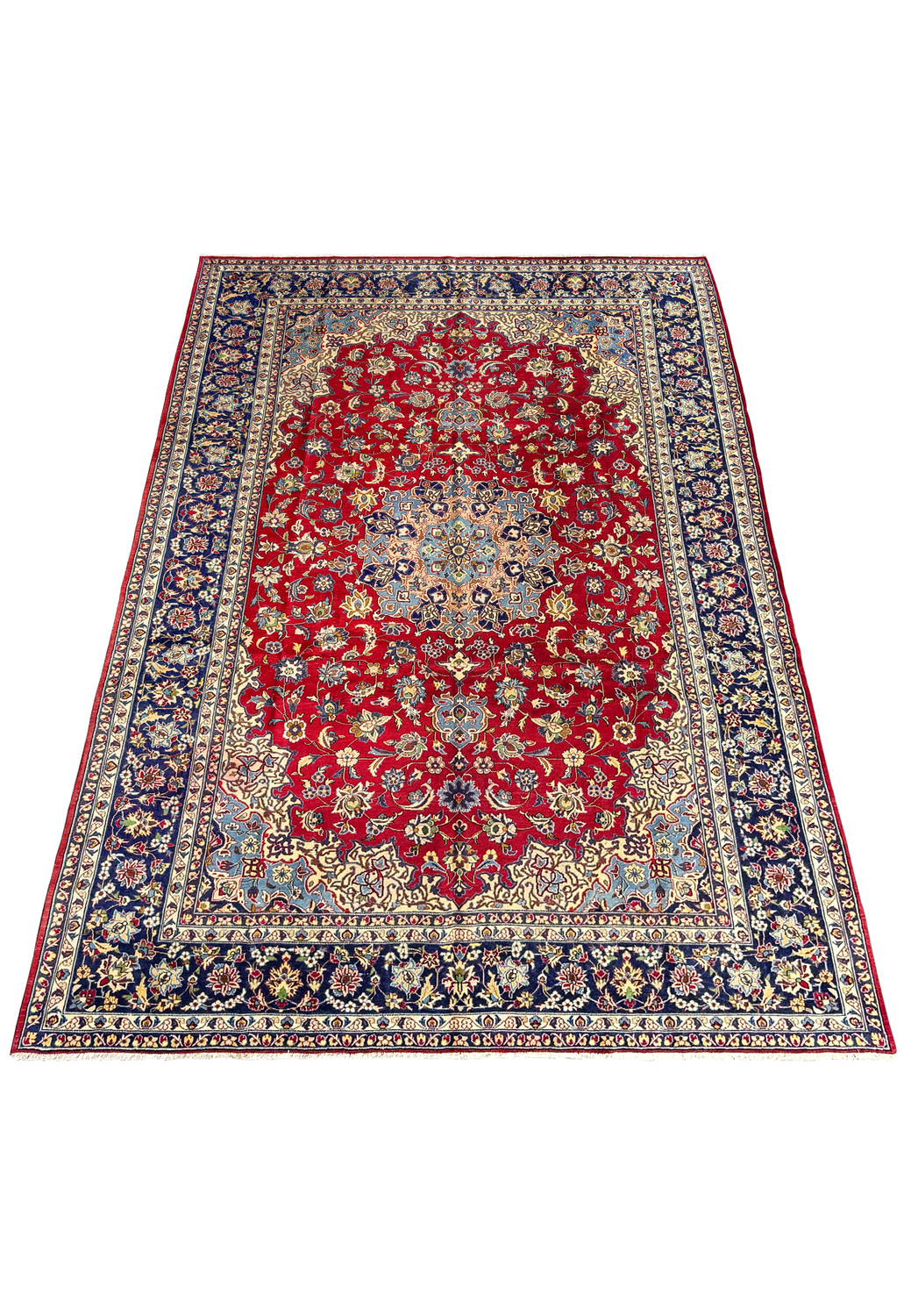 A full view of a large Persian Isfahan rug laid out on a flat surface, showcasing the rich red field and the dense, complex patterns in shades of blue, gold, and cream, bordered by navy blue edges