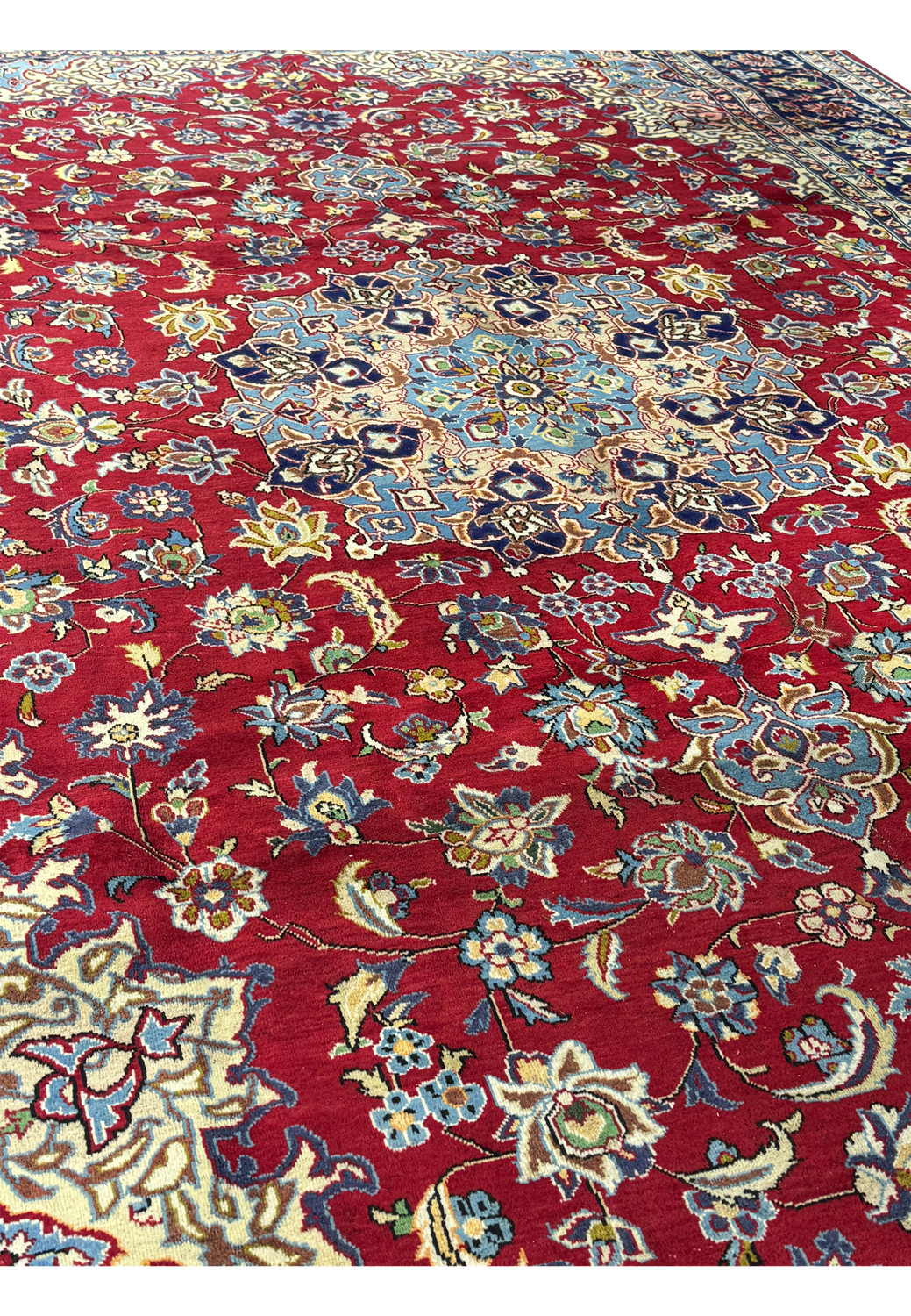 Angled view of a 10 x 15'6" Persian Isfahan rug, featuring a crimson field with a blue central medallion, ornate floral motifs, and a dark navy border