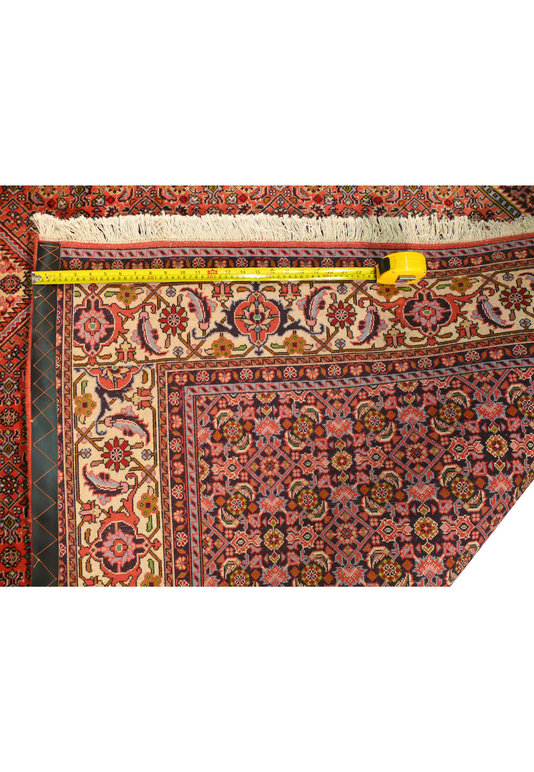 The backside of the Persian Bijar rug, showing the reverse patterns that mirror the front, indicating the high density and precision of the weave, with fringes lining the edges and a wooden rod supporting the rug's display.