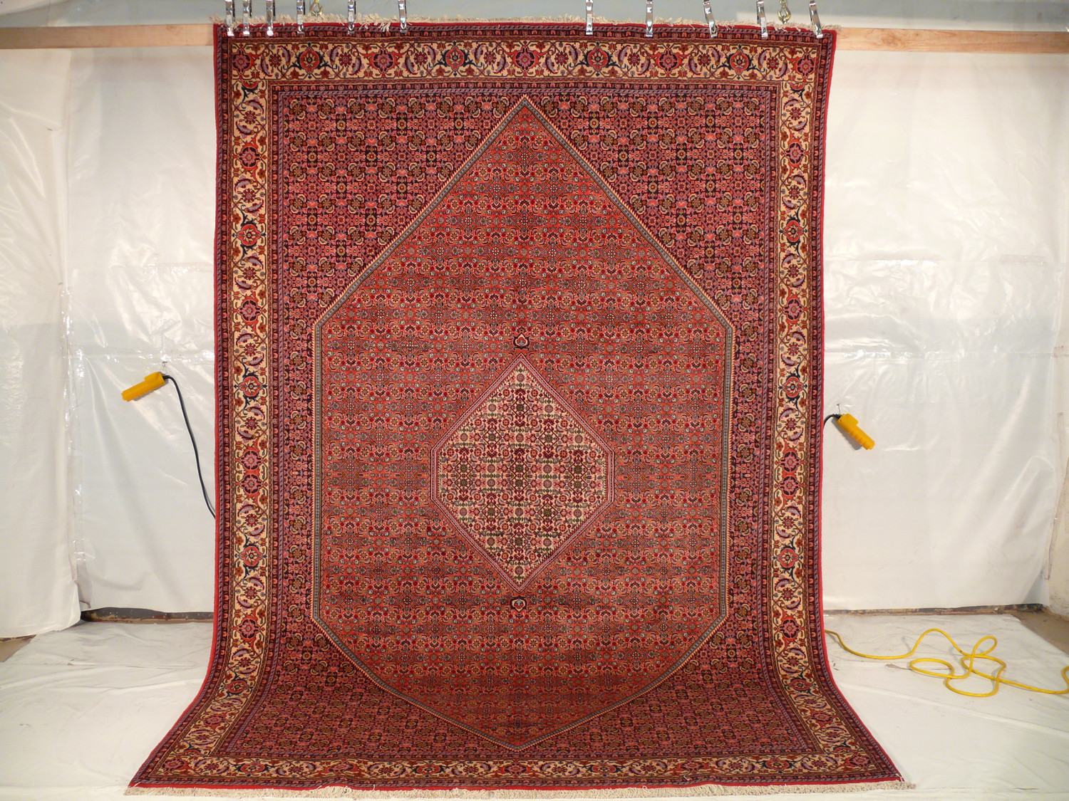 The Persian Bijar Iron Rug is shown hanging vertically, revealing its full splendor and the gravity-defying thickness of its weave, with its patterns cascading down its length, surrounded by a makeshift white backdrop and yellow measuring tape on the floor.