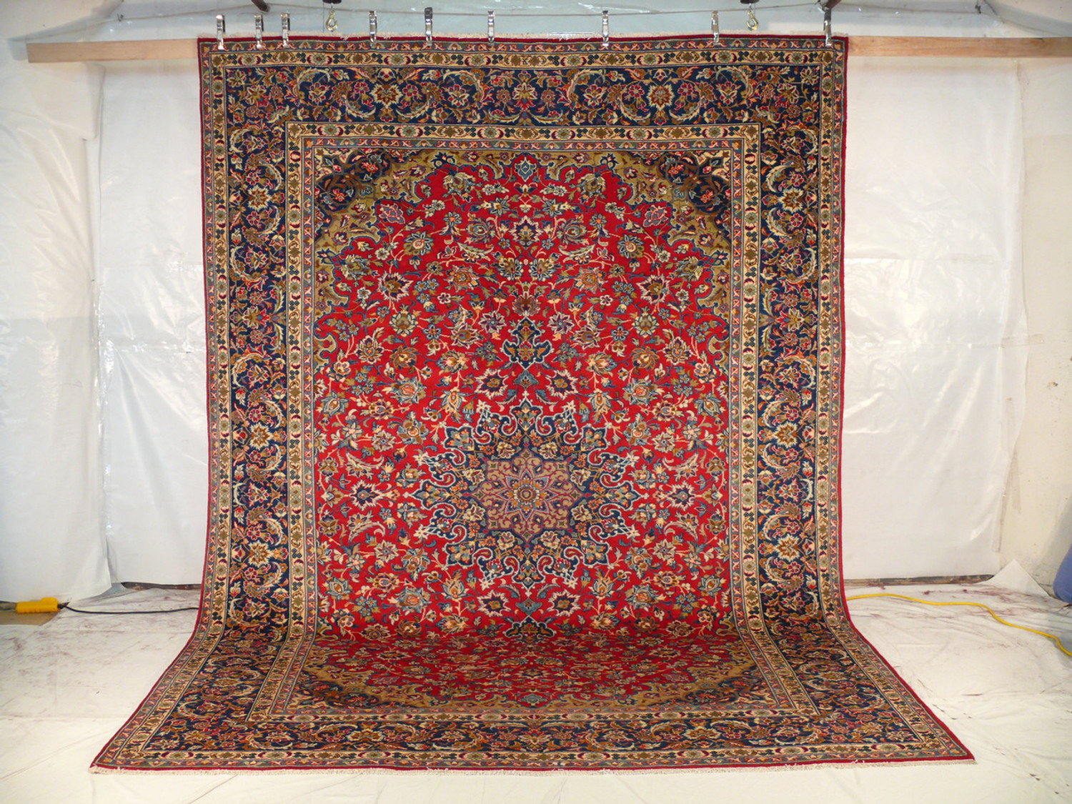 Full-length view of the 10x13 Persian Isfahan rug suspended vertically, exhibiting its grand scale, with the rich reds and deep blues of the field and border vividly on display against a neutral backdrop