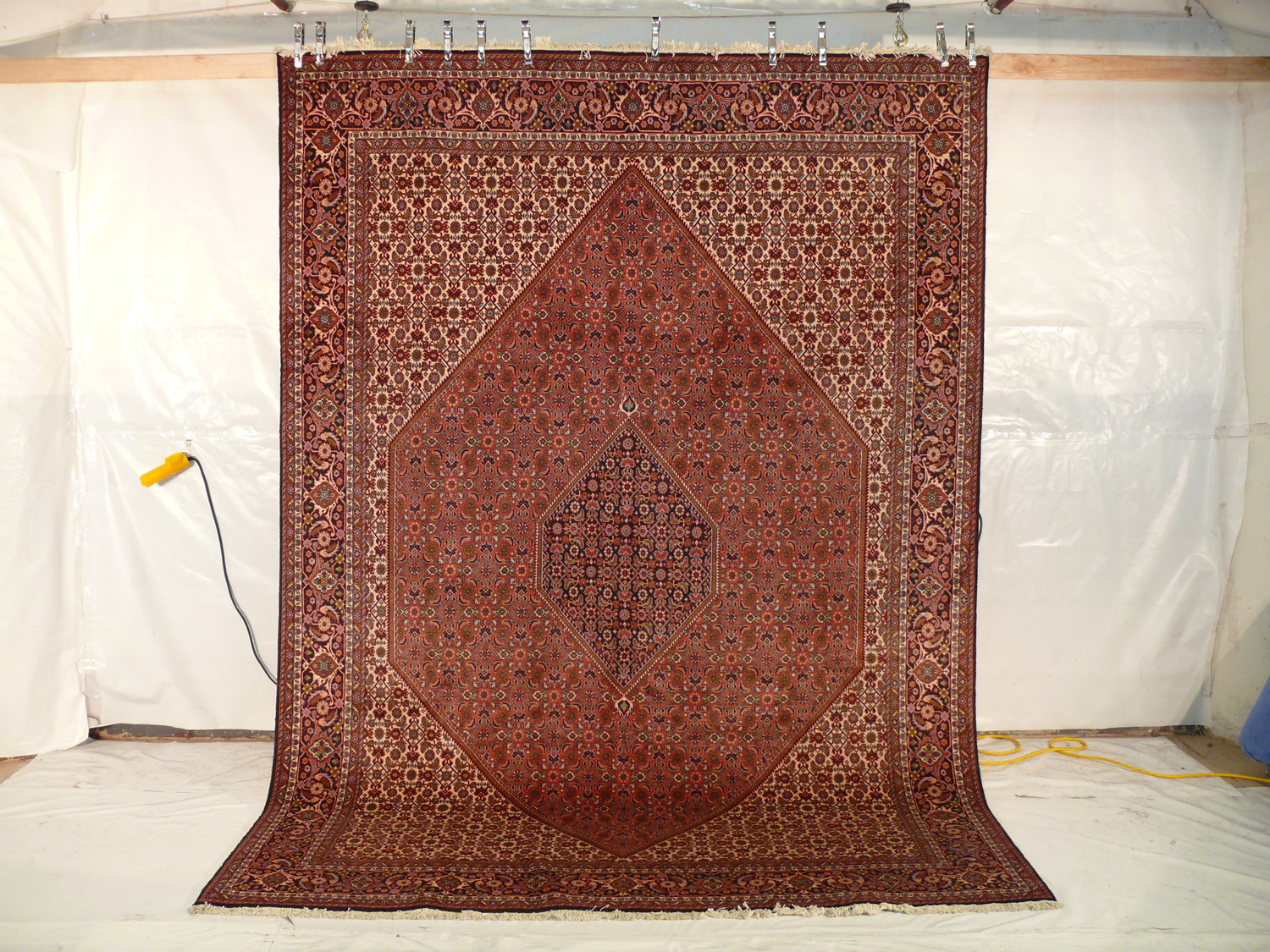 The Persian Bijar Iron Rug displayed hanging, revealing the full design with a prominent central medallion and densely patterned field, set against a white backdrop with a yellow extension cord on the floor.