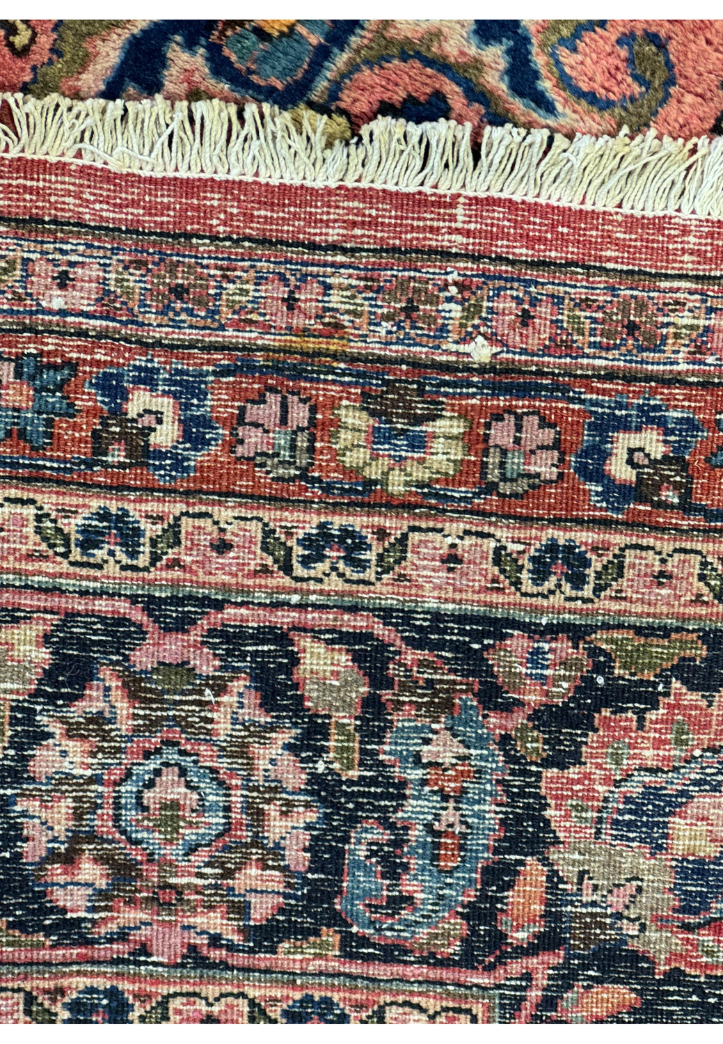 Detailed image of the fringe and border of a Persian Mashad rug. The image displays the fine white fringe, which transitions into the intricate border pattern featuring geometric and floral motifs in a rich array of colors such as deep blue, red, and pink on a dark navy background