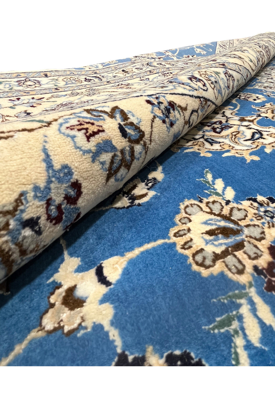 Hand-knotted Persian Nain rug partially rolled showing blue field and beige floral border.