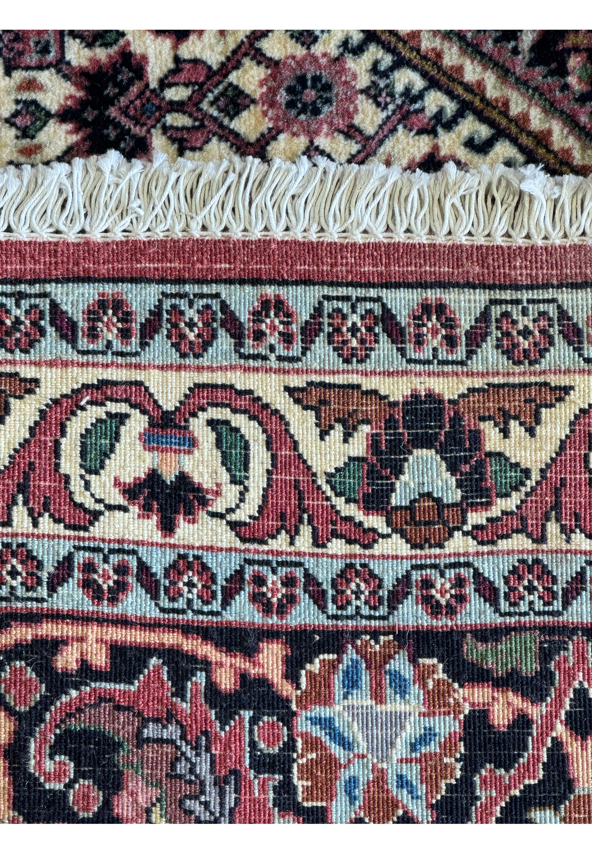 Close-up of a rolled Bijar Persian rug showcasing intricate patterns and vibrant colors. The rug features a dense weave with floral and geometric motifs in shades of red, blue, orange, and black on a pinkish-red background, with a soft, plush pile that offers a luxurious texture.