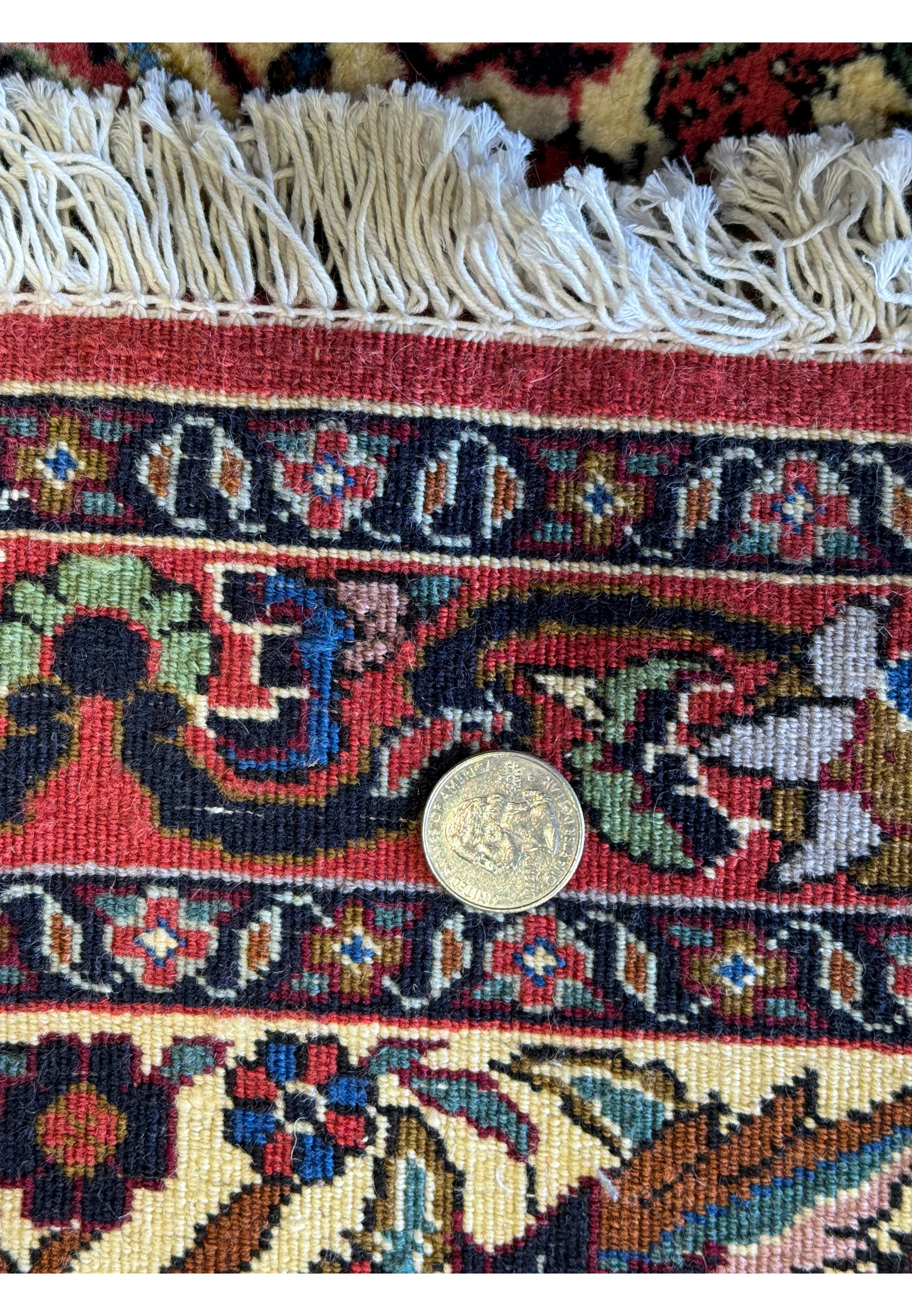 A close-up view of the rug’s fringe and detailed edge patterns with a quarter placed for scale, displaying the fine craftsmanship and scale of the motifs.