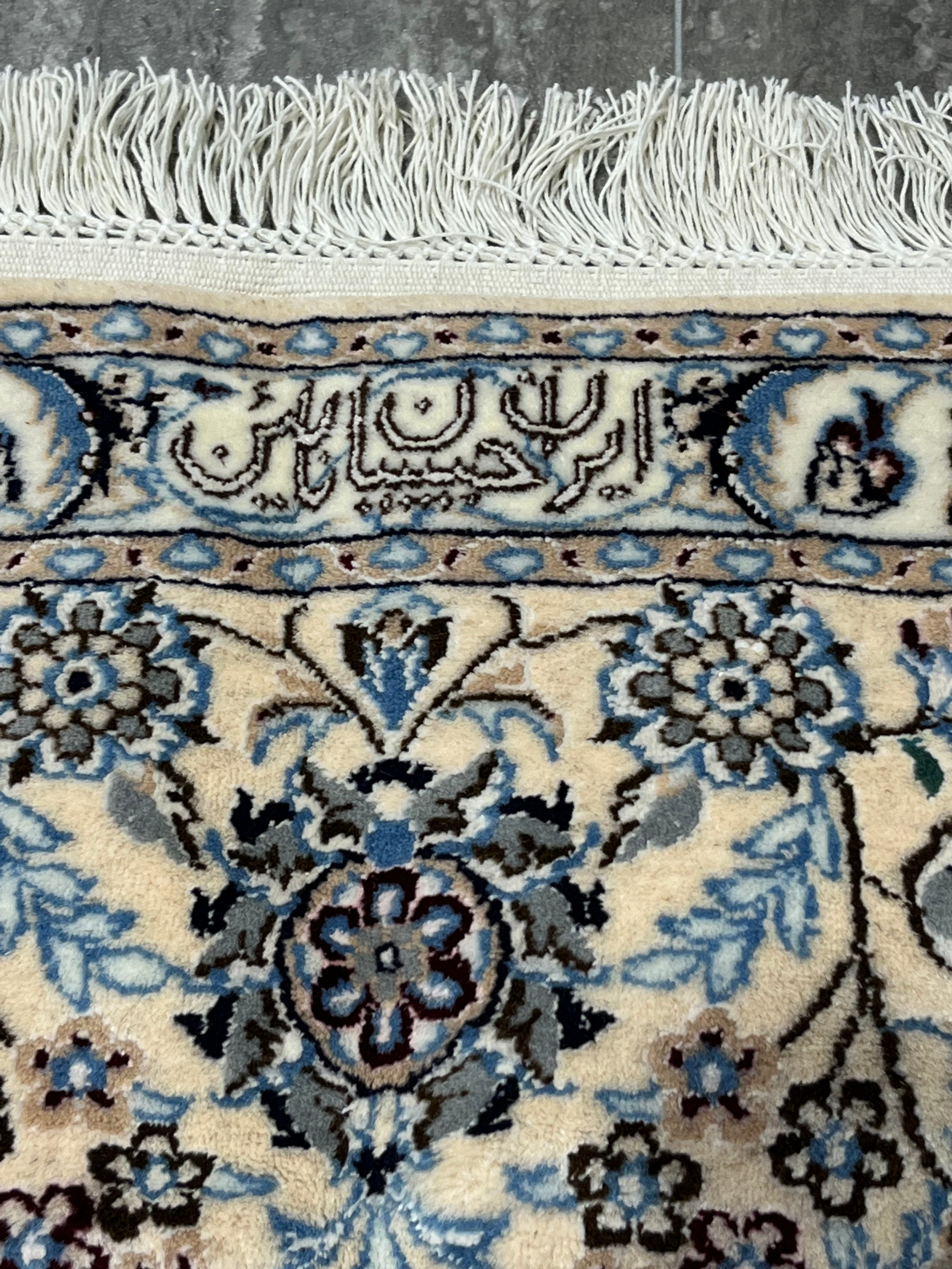Close-up of Persian Nain 6LA Habibian signature woven into the border, showing fine wool and silk details, ivory ground, soft blue floral motifs, and hand-finished fringe