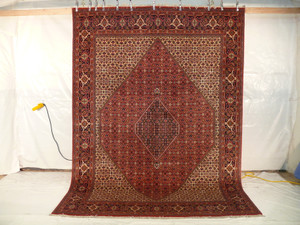 The Persian Bijar Iron Rug hanging vertically, revealing its full size and the dramatic impact of its traditional design against a plain white backdrop, with a yellow extension cord lying on the floor.