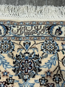 Close-up of Persian Nain 6LA Habibian signature woven into the border, showing fine wool and silk details, ivory ground, soft blue floral motifs, and hand-finished fringe