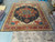 12' x 15' Serapi oriental rug displayed flat on showroom floor beside rolled inventory rugs