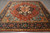 Angled view of coral red Serapi Heriz square rug with navy main border and ivory fringe