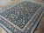 Angled view showing dark blue field, sage green border and dense hand-knotted wool pile