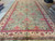 Angled view of green wool rug showing ivory main border with palmettes and pink rosettes