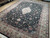 Angled showroom view showing dense lustrous wool pile, black field and wide ivory floral border