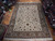 Ivory Tabriz rug laid on parquet showroom floor beside rolled Persian rugs, tan floral border visible