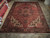 Semi-antique Heriz rug displayed on showroom tile floor beside stacked Persian runners and folded rugs