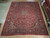 Large 11x14 Kashan rug displayed on wood floor beside rolled Persian rugs in the showroom