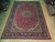 Red hand-knotted wool Tabriz rug laid flat on light wood showroom floor beside rolled rugs