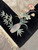 Carved vase holding lotus blossoms and bamboo above hand tied ivory fringe on black corner