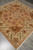 Angled view of hand-knotted wool square rug with cream floral border and ivory cotton fringe