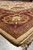 Angled corner view showing burgundy border palmette, taupe guard band and thick wool pile