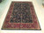 Hand-knotted wool rug displayed on light wood floor showing black field and deep burgundy border