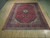 Red Kashan area rug shown full length on pale wood floor with dark border and fringe