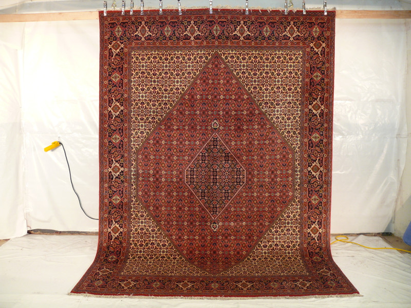 The Persian Bijar Iron Rug hanging vertically, revealing its full size and the dramatic impact of its traditional design against a plain white backdrop, with a yellow extension cord lying on the floor.