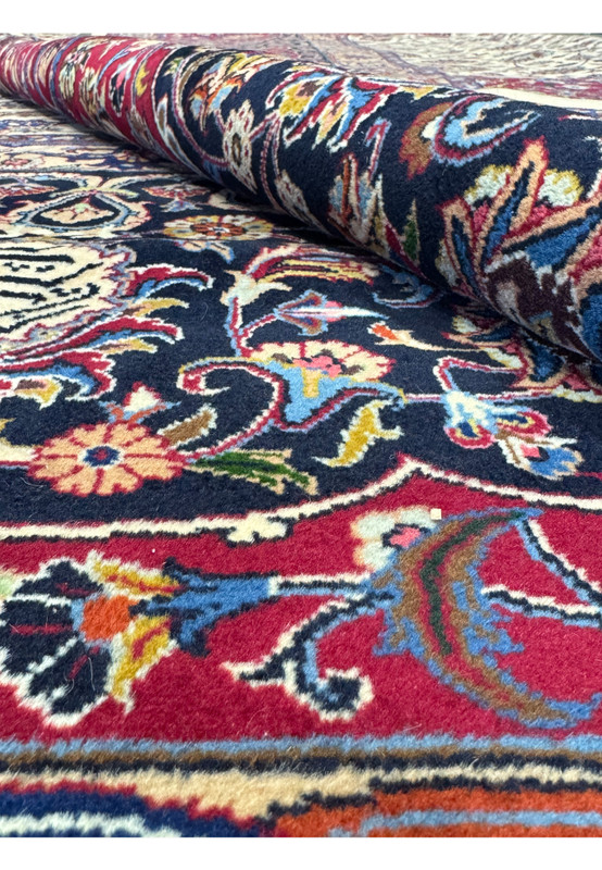 Close-up view of a Persian Mashad rug partially rolled, highlighting vibrant floral patterns and intricate motifs in deep red, blue, and ivory, showcasing the rich colors and detailed craftsmanship typical of traditional Persian rugs