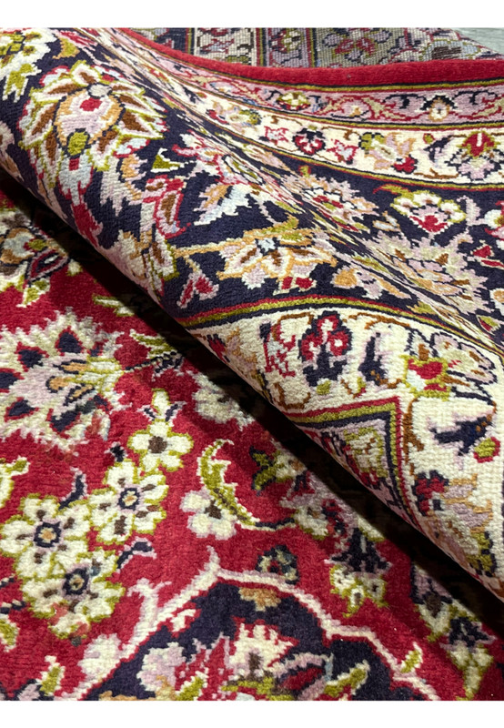 Rolled edge of red Persian Isfahan rug displaying dense floral motifs, navy border detailing, and finely woven wool pile.