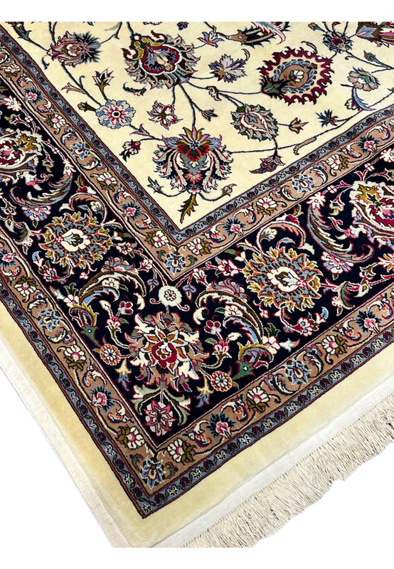 Angled view of signed Persian Mashad Saber rug showing ivory floral field and dark navy botanical border with fine wool detailing