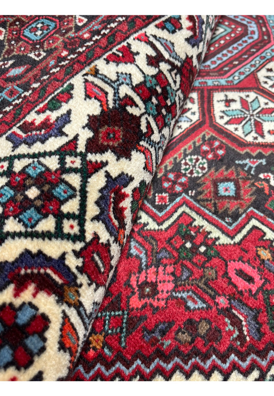 Rolled corner view of a 3'4 x 5'5 Persian Gholtogh rug revealing thick wool pile, sharp tribal pattern definition, rich red ground, ivory detailing, and turquoise and navy accent colors