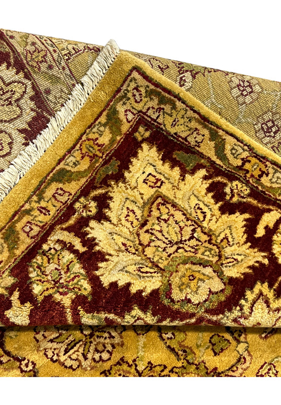 Folded edge of gold and burgundy Indian Agra rug showing wool pile thickness, fringe detail and tightly knotted construction.