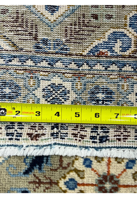 Extreme close-up of a Persian Tabriz rug with a measuring tape showing knot density, revealing tightly packed individual knots in the border area with blue rosettes and brown vine details on the fine cotton foundation.