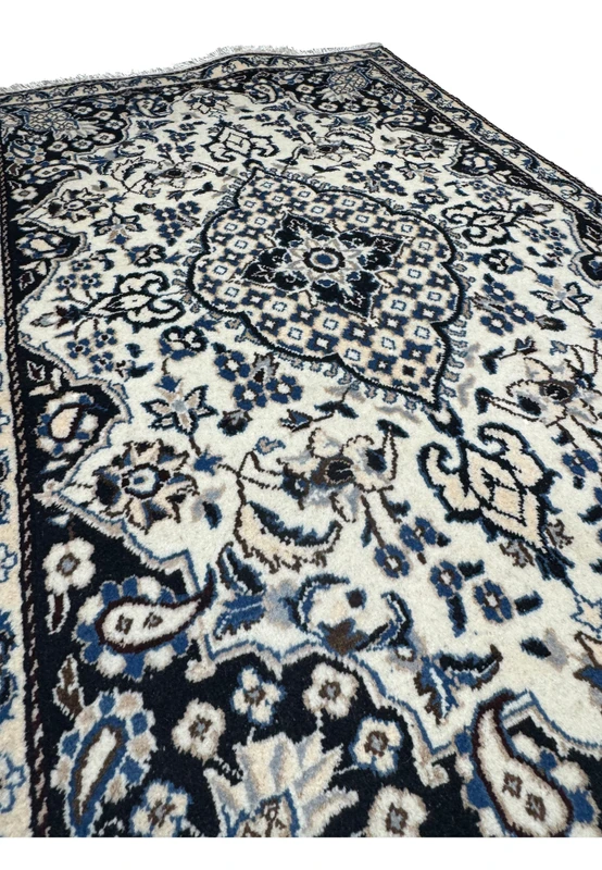 Angled studio view of Persian Nain rug from the foot end, layered floral medallion glowing against ivory and navy palette