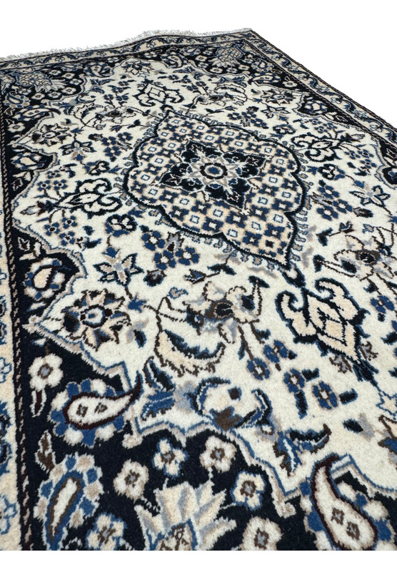 Angled studio view of Persian Nain rug from the foot end, layered floral medallion glowing against ivory and navy palette