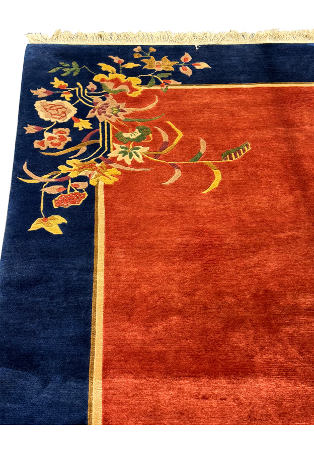Perspective view of the modern Oriental Art Deco rug lying flat, displaying the lush burnt orange center and navy border with a corner floral motif.