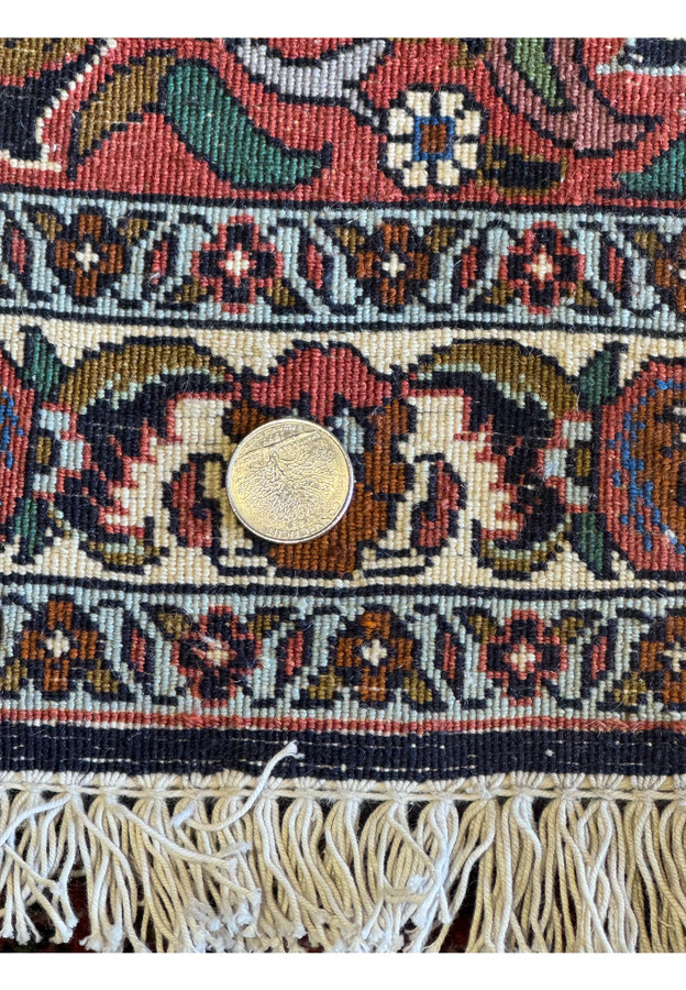 A close view of the Bijar rug's edge, featuring a coin for scale, showing the tight weave and elaborate border design