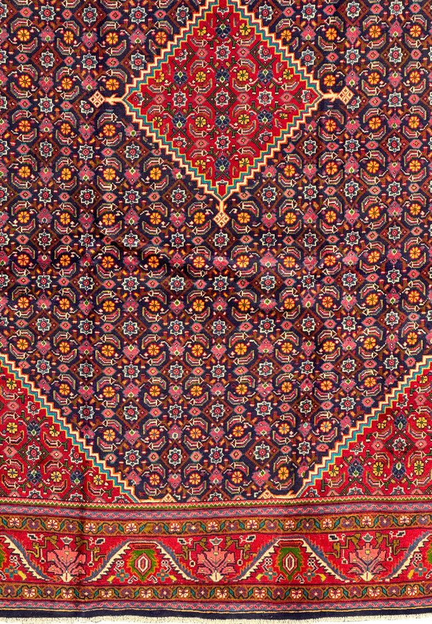 Angled shot of a Persian Mahal Rug, focusing on the detailed border and the harmony of the overall design and color scheme. The vibrant red border with its intricate geometric patterns frames the navy central field adorned with floral motifs. This perspective highlights the craftsmanship and the luxurious texture of the rug, making it a visually striking piece that enhances any room's decor.