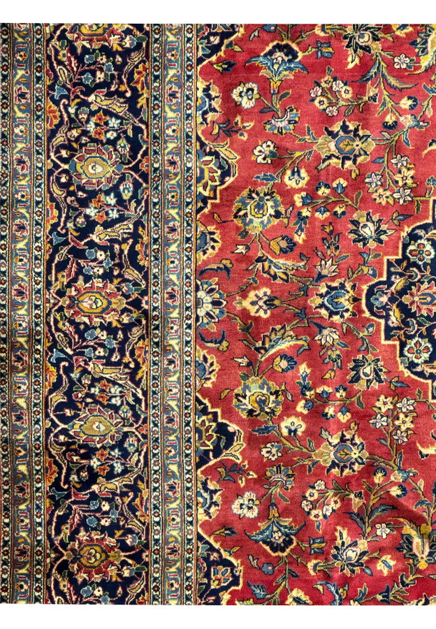 Partial view of a Persian Kashan rug, focusing on the transition from the central field to the border. The red field features intricate floral designs, leading to a multi-layered border with harmonious color combinations of blue, gold, and ivory. The craftsmanship is evident in the precise knotting and detailed patterns, making this rug a beautiful and durable addition to any home decor.