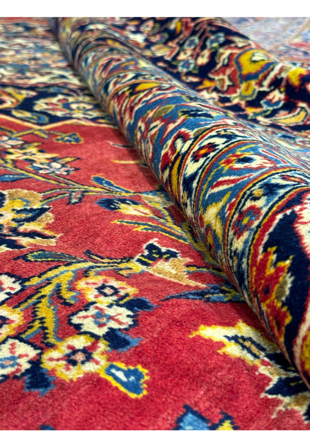 Perspective view of a Persian Kashan rug, showing the luxurious texture and detailed design. The rug is partially rolled to reveal the intricate border and floral patterns against a red field. The rich colors and high-quality materials are evident, making this rug a stunning addition to any room. This image emphasizes the durability and elegance of Kashan rugs.