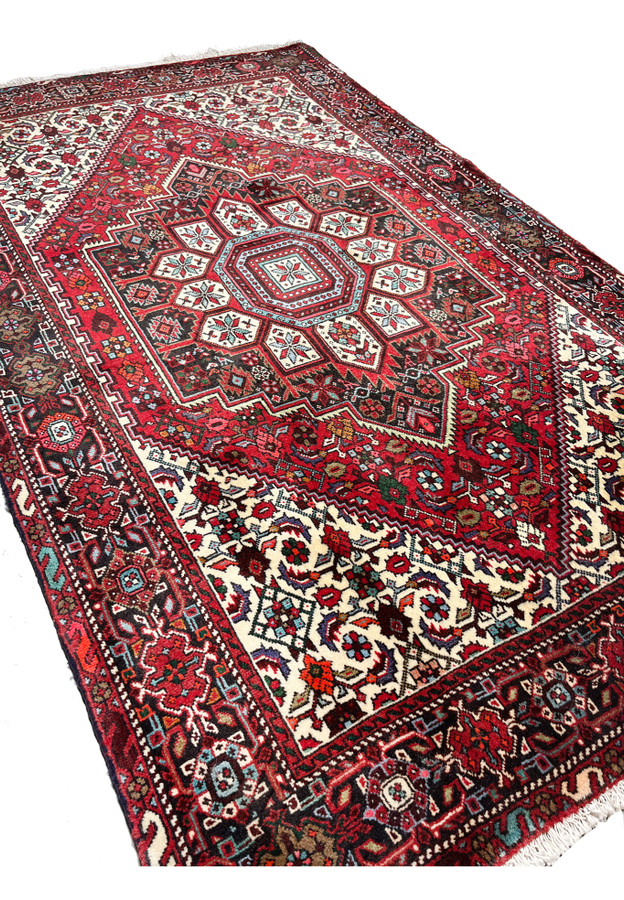 Angled view of a Persian Gholtogh rug emphasizing pile height, saturated red and navy colors, bold medallion presence, and sturdy tribal build