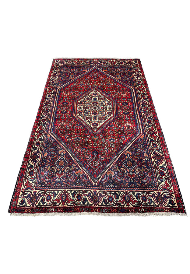 Hand-knotted Persian Gholtogh rug laid flat showing strong tribal symmetry, deep red background, ivory medallion, and dense village weaving.