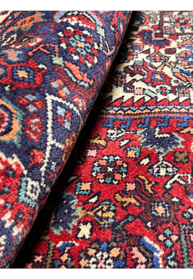 Rolled Persian Gholtogh rug revealing plush wool texture, saturated red field, navy blue accents, and finely detailed tribal motifs